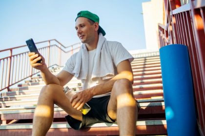 Fitness sport man sitting on stairs using mobile phone app relaxing post-workout in sunset light sha